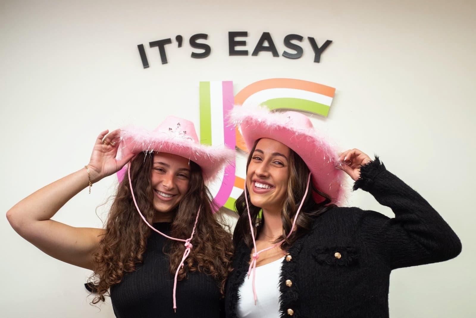 Two Just Start LA coaches in pink cowboy hats laughing in front of the It's Easy wall