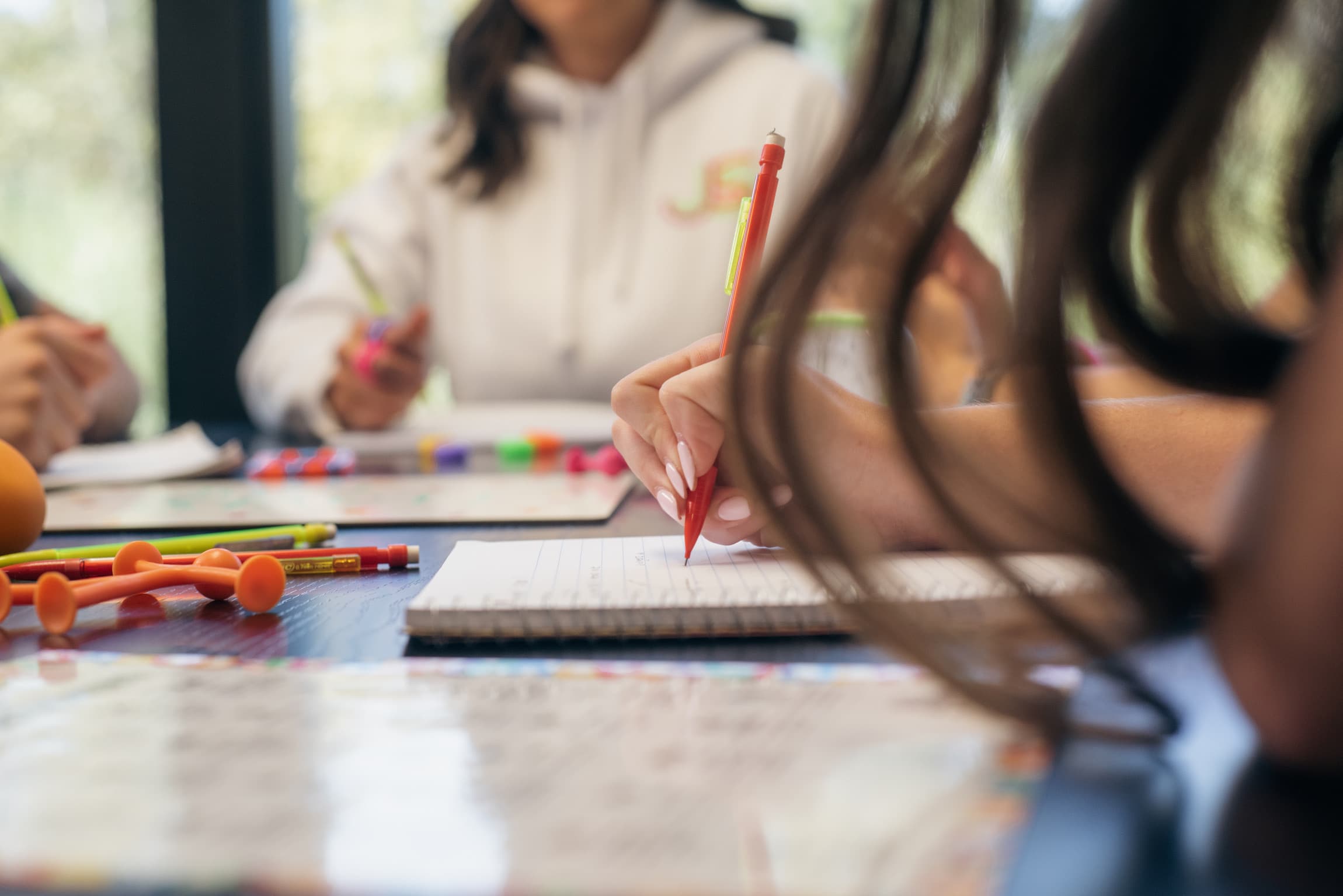 Students writing in notebooks at a shared table, with colorful pens and fidget tools spread across it