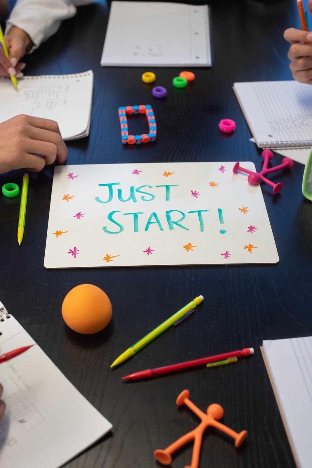 A session table covered with notebooks, colorful pens and fidget tools around a whiteboard reading Just Start!